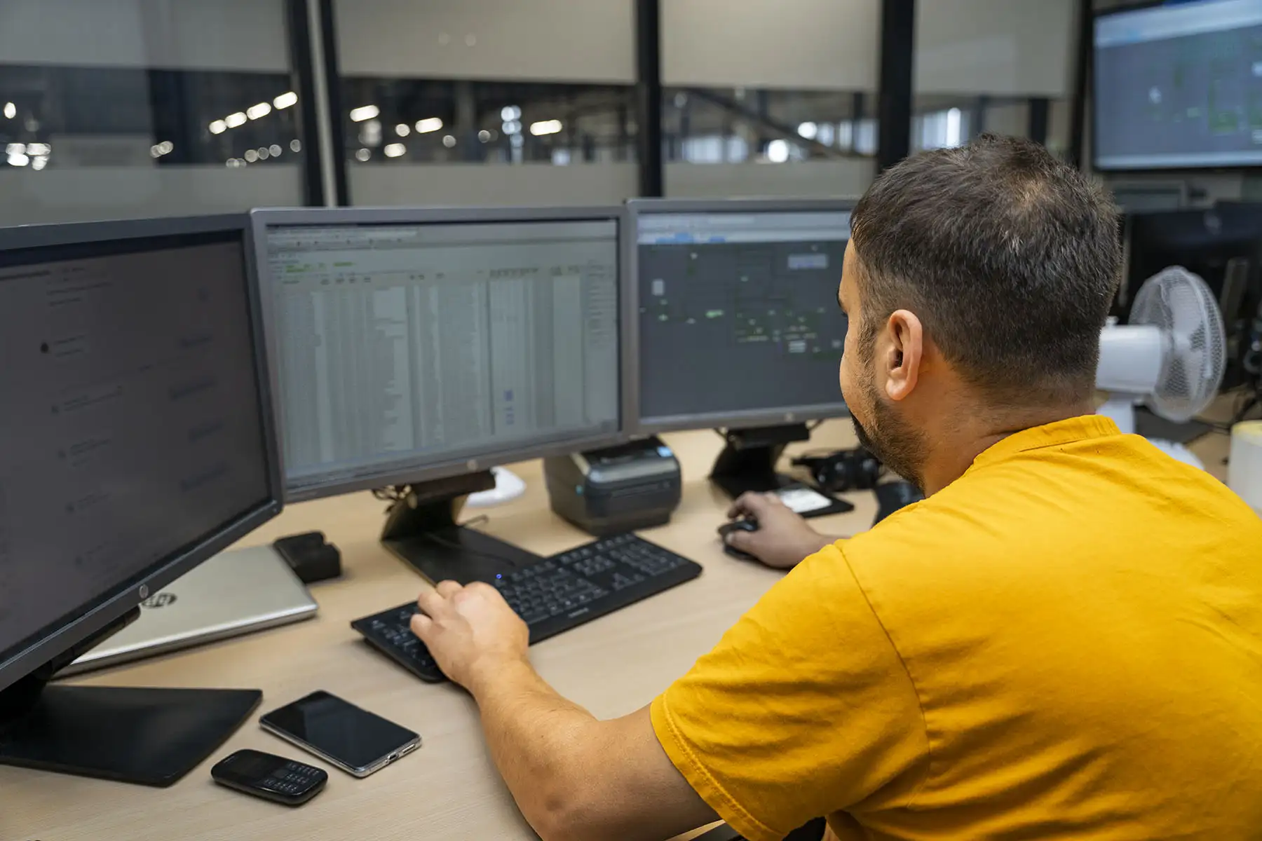 man in geel t-shirt achter een computer met 3 beeldschermen